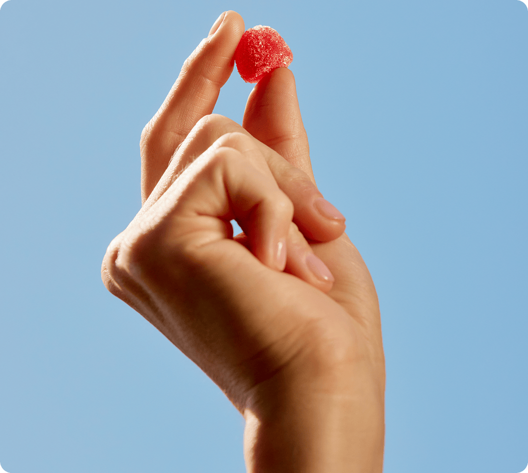 A hand holding a single red Needed prenatal gummy against a clear blue sky.