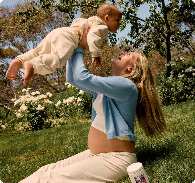 A pregnant woman sitting on the grass lifting a baby into the air