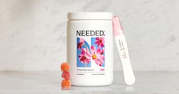 A jar of Needed Prenatal Multivitamin Gummy on a marble counter with a stack of gummies and a positive pregnancy test