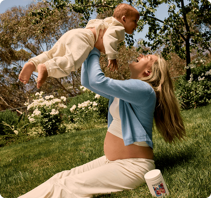 A mother lifting her baby in a garden, a jar of Needed Prenatal Multivitamin Gummy beside her