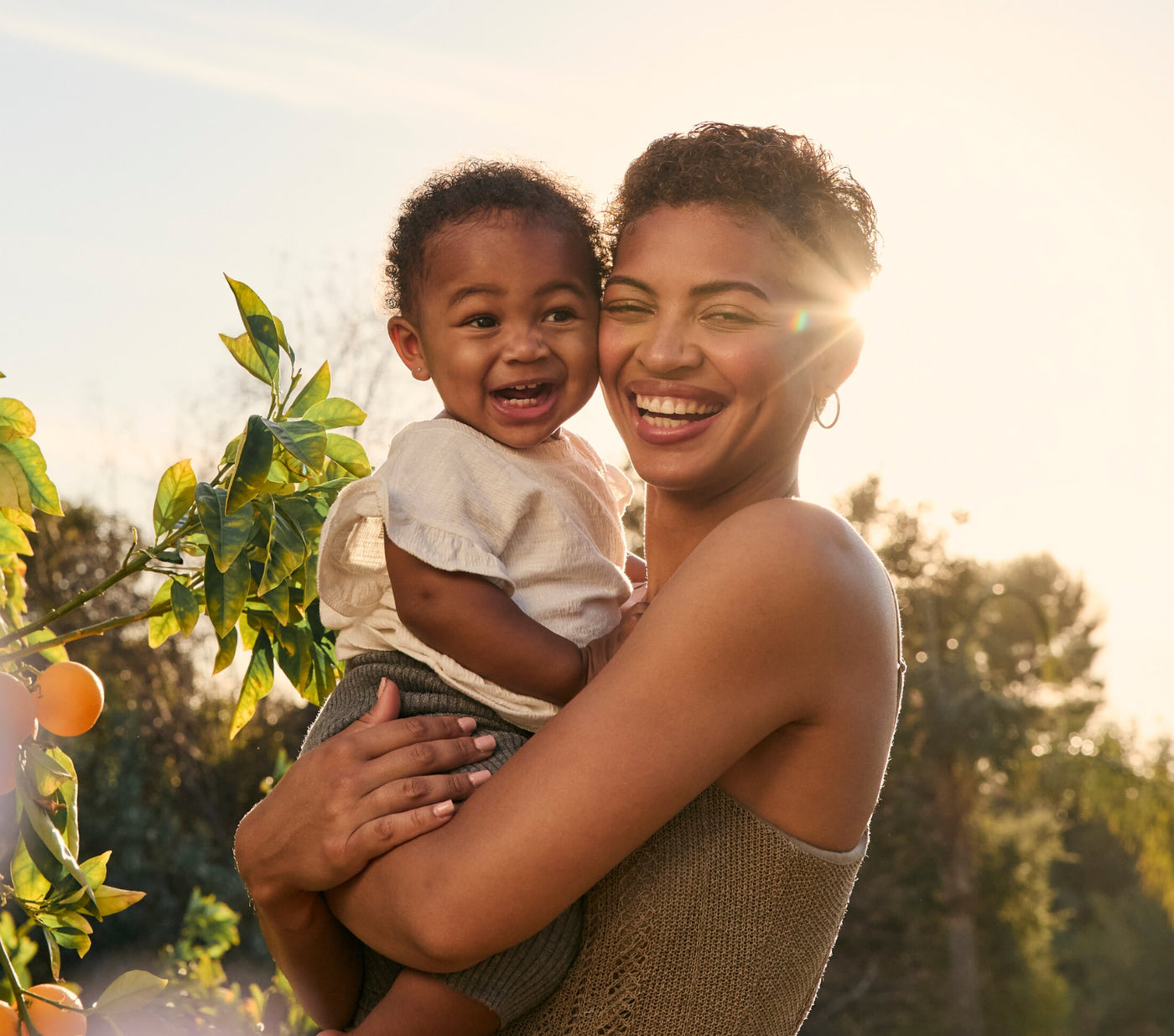 A smiling mother holding her baby in a sunlit orange grove